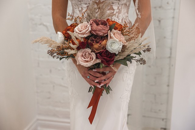 A shot of the bouquet, with fall coloured flowers, held by the bride in her wedding gown with white brick walls behind