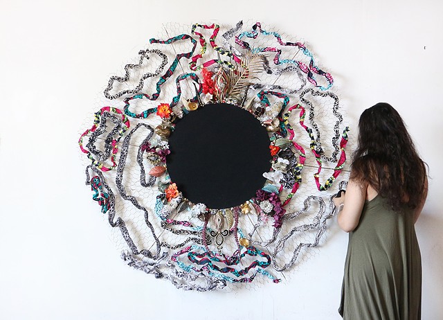 Circular wall sculpture, a deep black disk surrounded by radiating chicken wire woven with fabric and assemblage elements surrounding the inner black disk with brightly colored fabric flowers, beads, and ceramic objects