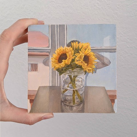 Hand holding miniature painting with still life of sunflowers in vase on table, figure peering in through window in background
