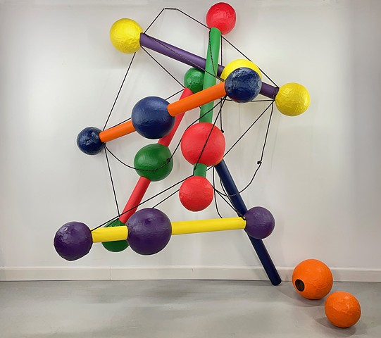 A large colorful sculpture that looks like a Manhatan tension toy. Sticks of red, blue, yellow, orange and purple are capped by paper mache balls. Unlike the original toy held together by elastics, this wall0mounted sculptural version is collapsing, droop