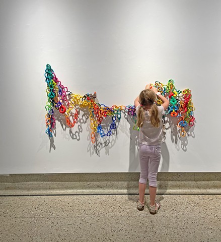 A colorful mass of infant linking rings creates an amorphous shape on the wall. A 6 year old child with long blonde pigtails, pink pants, and a white shirt is seen from the back, reconfiguring the rings.