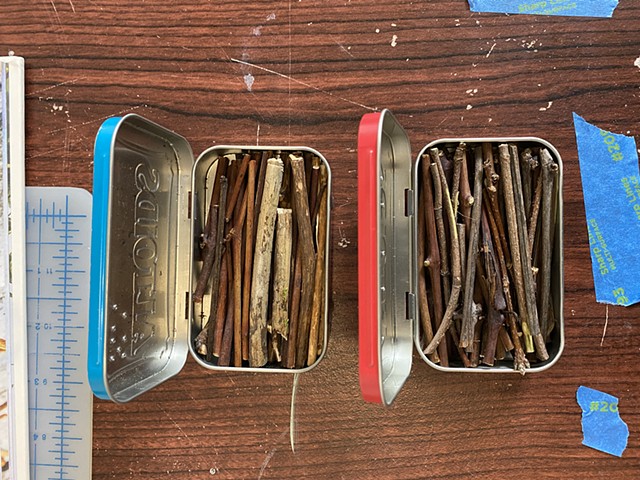 Pieces of dry wood have been cut to size and placed in metal containers with lids, which will be wrapped with wire to secure them. A single hole will be punched in the lid, allowing for hot air to escape when placed in the fire. 