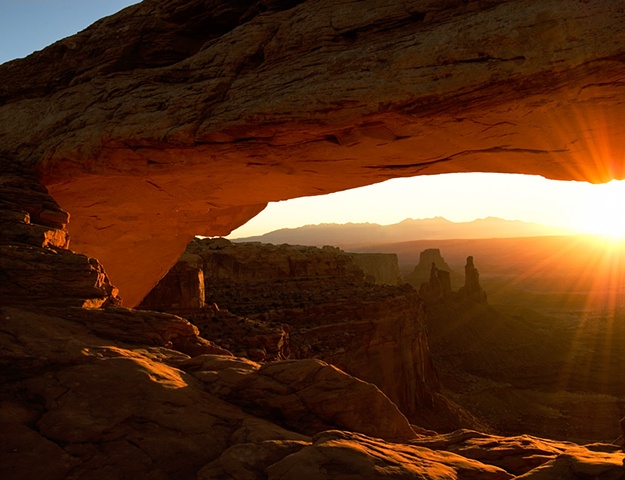 Mesa Arch sunrise, rays of the sun lighting the underside of the rock arch, and washer woman in the background