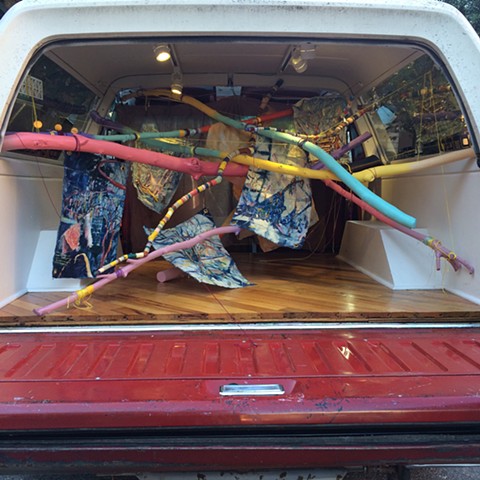 Brightly painted sticks crossing each other, providing hanging system for unstretched oil paintings on indigo dyed canvas, all installed in the back space of a 1991 Ford Bronco 