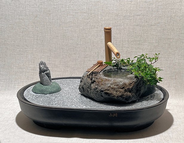 A tabletop version of a Japanese garden, with stone water basin, bamboo fountain, sentinel stone, and live plant.