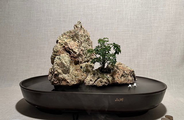 A tabletop fountain with deeply textured rock miniature islands, a tree, and two white cranes flying low through mist over the water