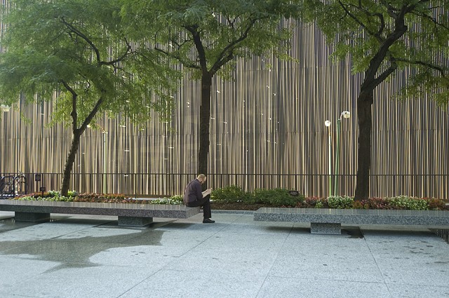 Man sits reading a book in a Michigan Avenue Plaza with the sun setting on a wall behind him that has a digital hum by Lucy Mueller