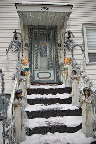 Dark and light skinned Christmas angels line the snow covered steps and porch of a house by Lucy Mueller