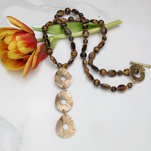 a trio of hammered brass radiant lined teardrop waves hang from a 28" strand of multiple shapes and sizes tiger's eye beads highlighted by brass spacer beads and finished with a hammered brass toggle #138
