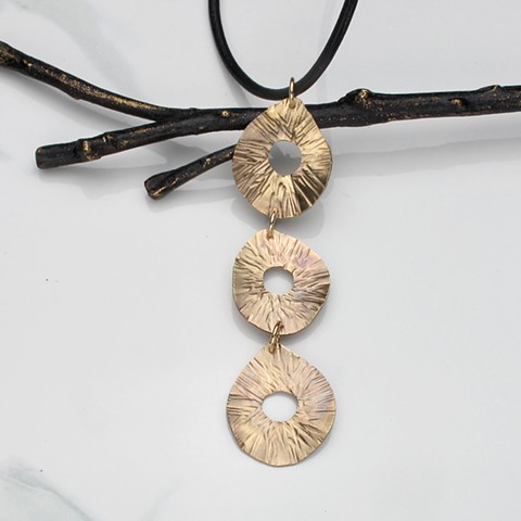 This hammered brass pendant features a trio of randiant lined waved teardrops on a 28" black leather cord, finished with a sterling silver hook & eye #137