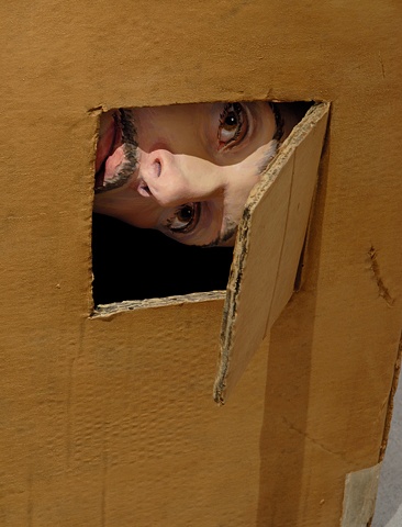 Sculpture of man in cardboard box- detail of face peeking out from cutout with flap