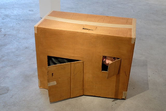 Figurative polychrome sculpture of adult man stuffed inside a cardboard box or child's playhouse