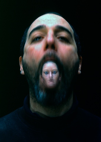 Blurry photograph of a man with a small sculpted, painted woman's head in his mouth
