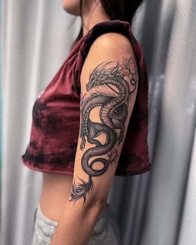 Strange world tattoo Calgary alberta Canada dragon tattoo in black and grey sketchy style with a couple of lotus style florals pieces in illustrative feminine dainty fineline soft upper arm tattoo from shoulder to elbow
