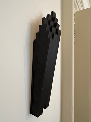 Contemporary sculpture exhibited on a wall. An abstract, geometric relief made of wood shims covered in a matte coating of black oil pastel. 