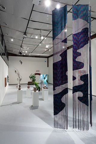 Three long woven panels hang installed in a two-story warehouse space. The woven sections line up between panels, mimicking an oil slick on water both in form and patterning.