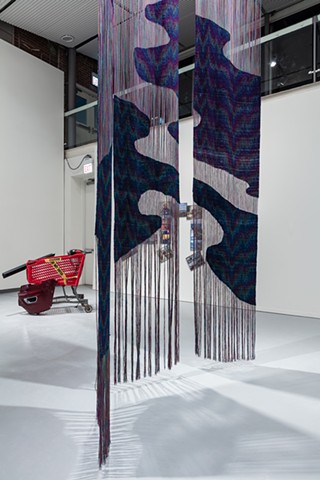 Three long woven panels hang installed in a two-story warehouse space. The woven sections line up between panels, mimicking an oil slick on water both in form and patterning.