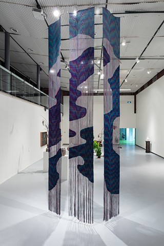 Three long woven panels hang installed in a two story warehouse space. The woven sections line up between panels, mimicking an oil slick on water both in form and patterning.