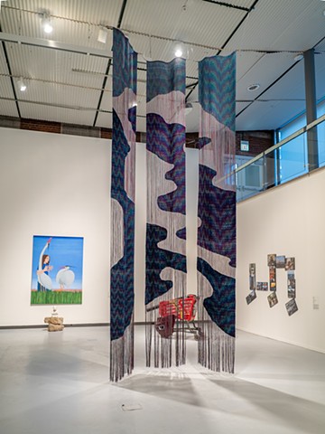 Three long woven panels hang installed in a two-story warehouse space. The woven sections line up between panels, mimicking an oil slick on water both in form and patterning.