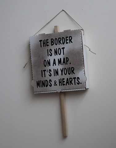 Robert Fields, "THE BORDER IS NOT ON A MAP." 2019. Adhesive vinyl, vegetable oil on corrugated paper board, with wood & twine. Speaks to refugees seeking asylum.
