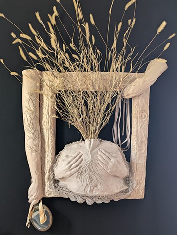 Cast paper sculpture composed of a frame, a bust with dried grasses growing from it, and hands