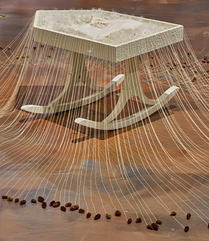 White cradle filled with milkweed seeds and an embroidered baby gown; surrounded by a halo of tiny pinecones on strings