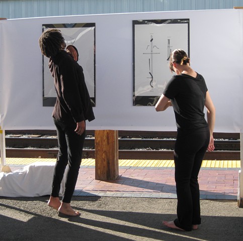 Two dancers in all black clothing dance in front of a graphic score, hanging outside in an arts festival. They interpret the vertically drawn shapes, some in heavy thick marks denoted downward pressure, and some in light marks denoting smaller gestures.