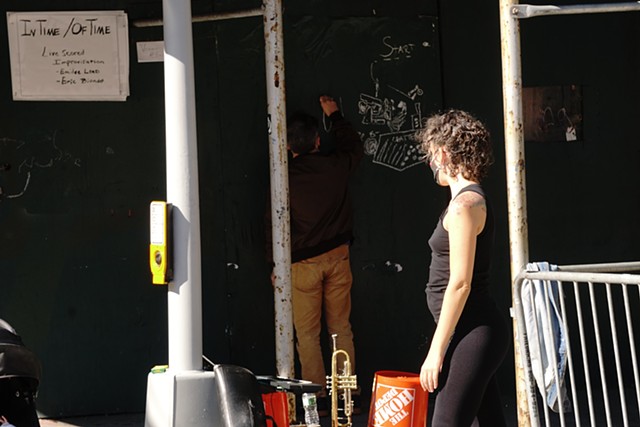 a man in light brown pants and dark brown jacket draws in chalk on a green wooden construction site wall at a street corner. A dancer, standing in the crosswalk in all black, watches him