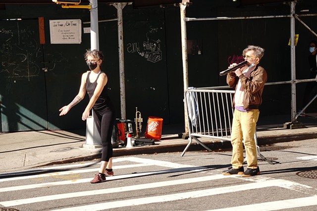 on a street corner and moving into the cross walk, a dancer all in black and wearing a black mask reaches forward with her palms open and up. Behind her a man in yellow pants and a brown jacket plays a Melodica.