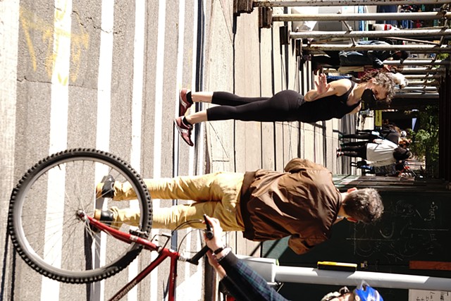 In the cross walk the dancer presses her right heel into the pavement in front of her left foot. Her right arm is bent up at the elbow and her right hands flicks open and behind her shoulder. A trumpet player in a brown jacket walks towards her. A cyclist