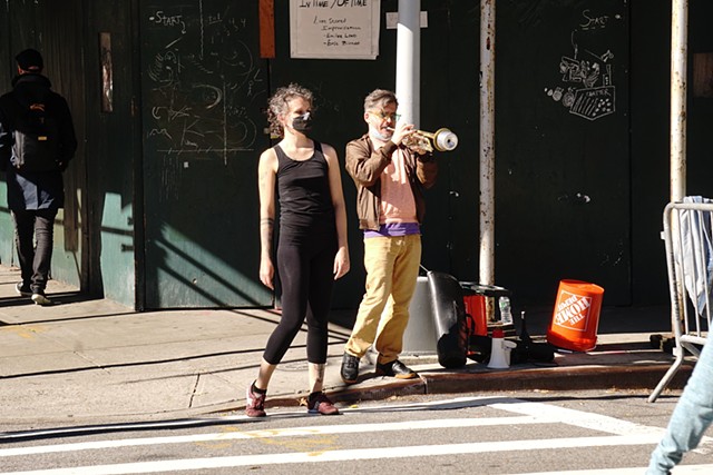 On a street corner, chalk drawings visible on a construction wall, stand a dancer and a musician playing the trumpet.
