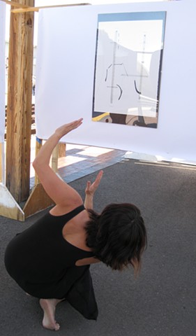 In front of a graphic score where only a couple dark half circle lines are visible because of the glare of the sun, a dancer interprets these marks. She is crouched and leaning into her right foot, her head far to the right over her center. Her left arm i