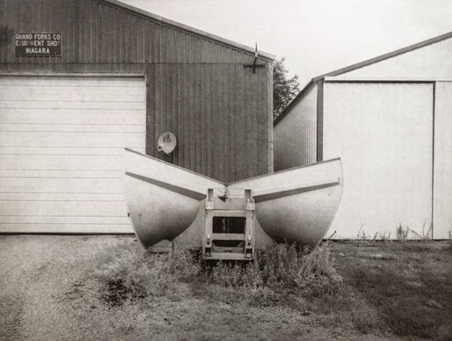 On a summer day a snowplow attachment sits in front of garage buildings in Niagara, North Dakota. One-plate polymer photogravure print on pescia paper by John Pearson.
