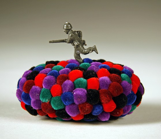 found rock covered in purple, red, turquoise, green & maroon pom poms with running toy army soldier on top by Holly Campbell