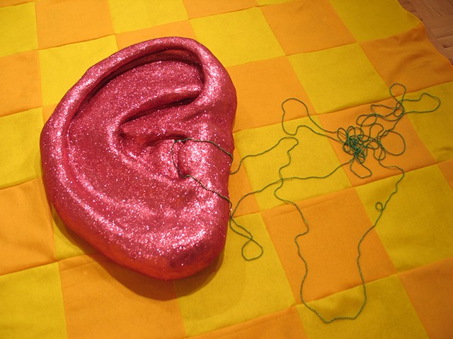 sculpture pink glittered ceramic ear with green strings of beads on checkerboard felt background by Holly Campbell