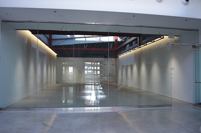 This is an image of an percievably empty gallery. The Gallery has three white walls and a glass front facing wall. On the walls you can kind of tell there are dark spots like there might be work on them, but nothing is hanging on the wall.