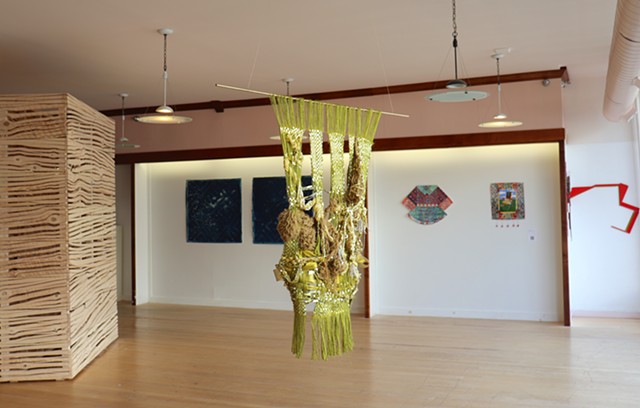 A gold and green tapestry is hanging from the ceiling in an exhibition space. Its green warp hangs off of a horizontal gold rod and further down are strands of shiny gold tinsel woven in checker patterns and split into four rows connected by knitted brown