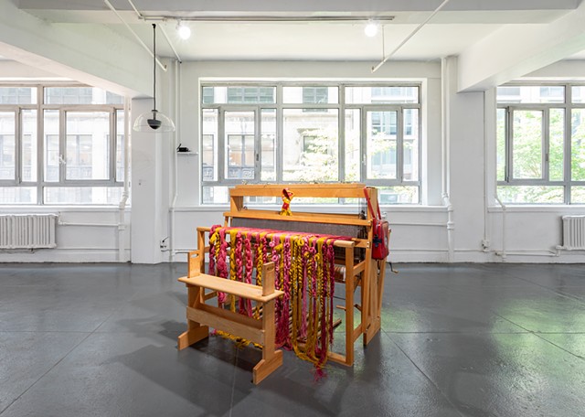 A loom with pinkish red and yellow yarn braids hang off in the middle of a gallery. 