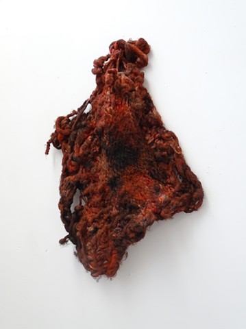 A knit wool sculpture hangs diagonally from one of its four cornes. The object is covered in oil paint with fleshy hues of red and black. The fabric is tightly knit in some areas and unravelling around the edges. 