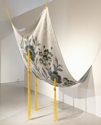 A blanket sculpture hangs from the ceiling in a white wall gallery.
