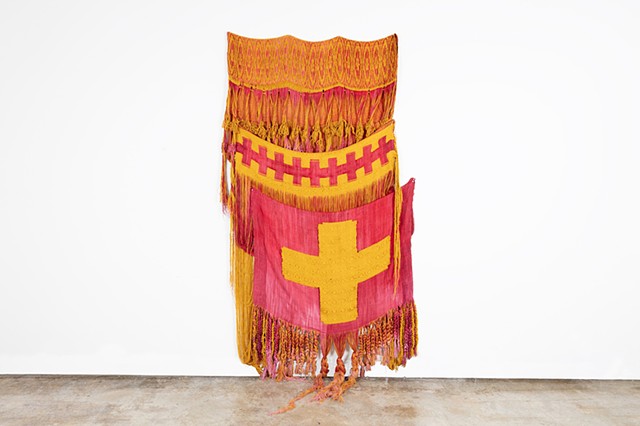 A large pink and yellow patterned textile hangs on a white wall. The patterns at the top are elongated circles of both colors. Then in the middle is a yellow foreground with a reddish line of connected crosses. At the bottom is a big section of a reddish background with a yellow balanced cross.