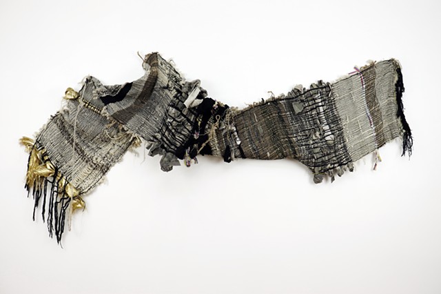 A textile hangs on a wall horizontally with one side flipped over. The textile is comprised of gold, wool, hemp, Colombian flags, camo print and fishnet in stripes and black fringe on each end. 