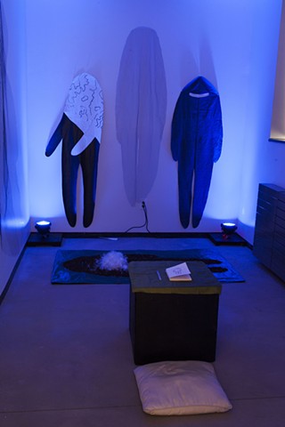 three cloth figures are pinned to the walls of a room illuminated by blue lights, there is a box in the center of the room with a small journal on top