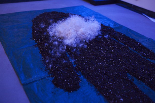 a figure made out of dirt and milkweed seeds lays on a tarp on the floor of a room illuminated by blue lights