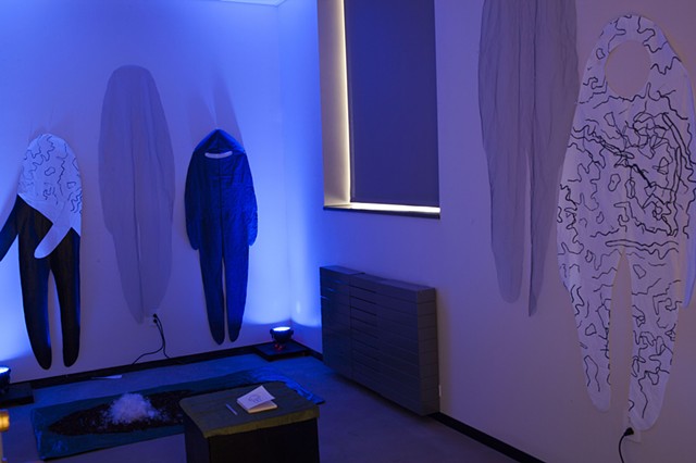 five cloth figures are pinned to the walls of a room illuminated by blue lights, there is a box in the center of the room with a small journal on top