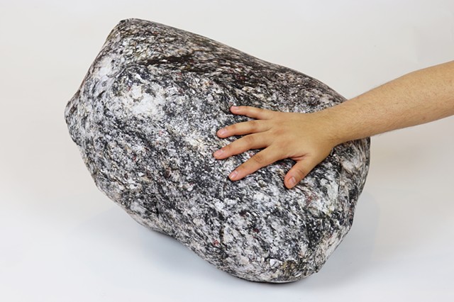  One rock pillow sits on a white backdrop. It’s roughly just under two feet in diameter and made from different shapes sewn together. It has realistic crevices and ever so slightly visible seams. This rock is primarily dark gray but is intricately speckle