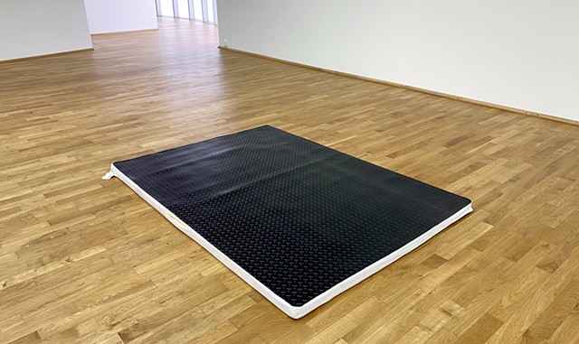 Resting in the middle of a wooden floor and surrounded by white museum walls, a mattress topper with a white fabric cover can be seen from underneath black diamond plate rubber flooring which has been cut to fit the dimensions of the mattress topper. The 