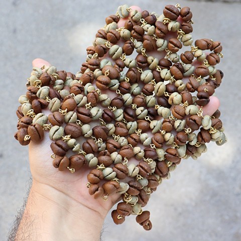 A mesh-like almost square made from a chain of green unroasted coffee beans woven like a fabric over and under through another chain of brown roasted coffee beans. The beans fit together in pairs and are connected by tiny loops of golden brass that go through the center of the beans like beads. Here I hold the piece in my hand, open palm face up, as the beads bunch together and the flexible mesh forms to my hand. 