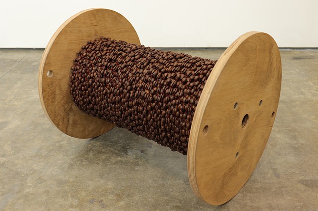 A wooden cable reel, which looks like an oversized spool, sits horizontally on it's side on a concrete floor in front of a white wall. The cylindrical middle of the cable reel is fully covered by layers of coffee beans, which have been glued together in p