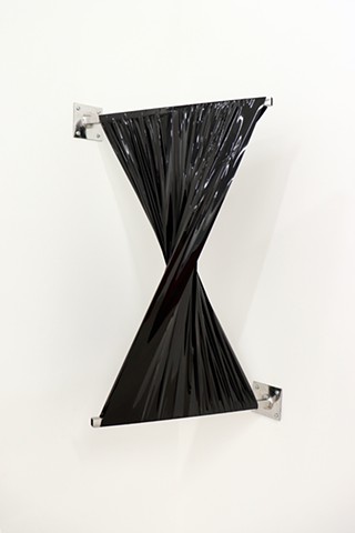 Strips of black shiny material wrap across two silver metal rods which extend outwards from a white wall and are anchored with square flanges. The piece is installed in a corner and the twist of the material creates a bow tie shape, with the middle of the